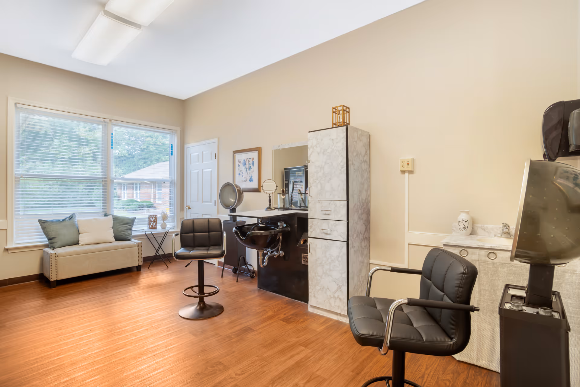 Bright salon/styling room with barber chairs, a shampoo basin, mirror and a cushioned bench by large windows.