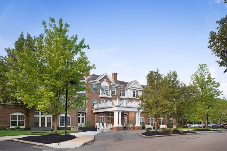 Exterior view of a multi-story brick senior living facility with white trim, a covered entrance supported by columns, several rocking chairs on the porch, surrounded by green trees and a parking area.