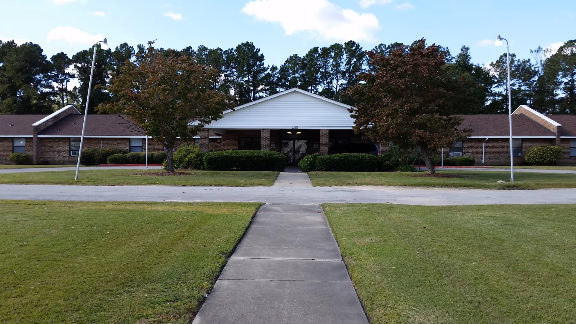 Front exterior view of a single-story brick building with a white gabled entrance, flanked by two trees and bushes, with a concrete walkway leading up to the entrance and street lamps on either side.