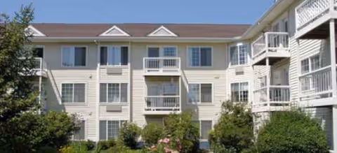 Three-story beige senior living building with balconies and landscaped shrubs in front