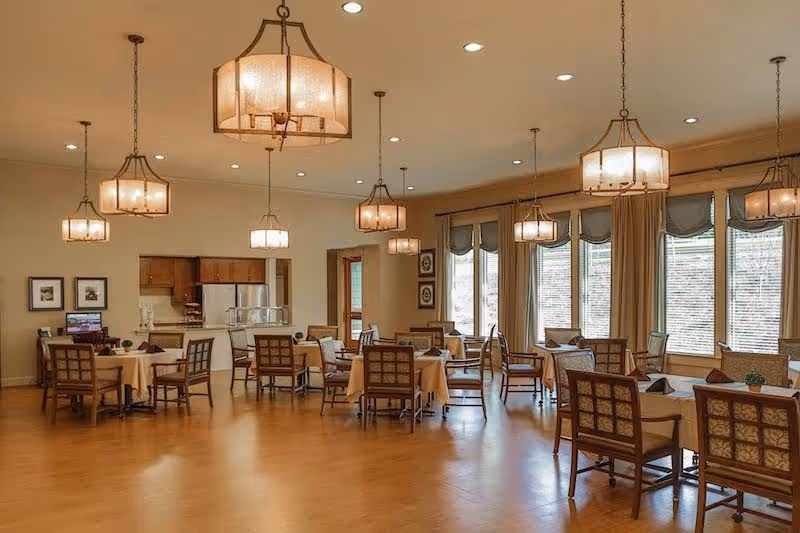 Spacious dining room with several set tables and chairs, large windows with curtains, and multiple hanging light fixtures.