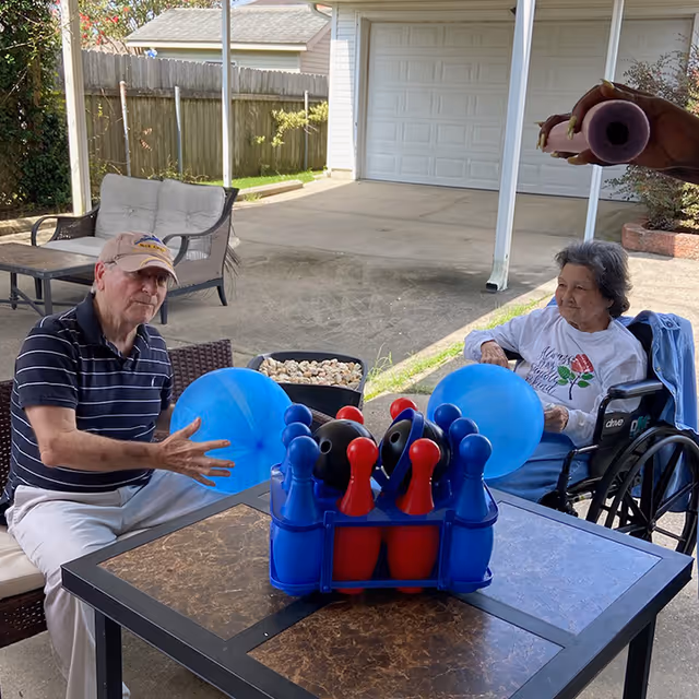 An elderly man and woman sitting outdoors under a covered patio. The man is seated on a bench holding a blue balloon, and the woman is in a wheelchair also holding a blue balloon. On the table in front of them is a bowling game set with red and blue pins and black balls. In the background, there is a garage door and a wooden fence.