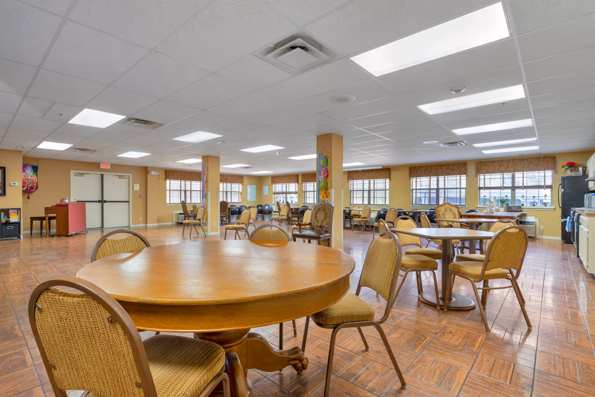 Spacious communal dining/activity room with round wooden tables and chairs, large windows, and a kitchenette area.