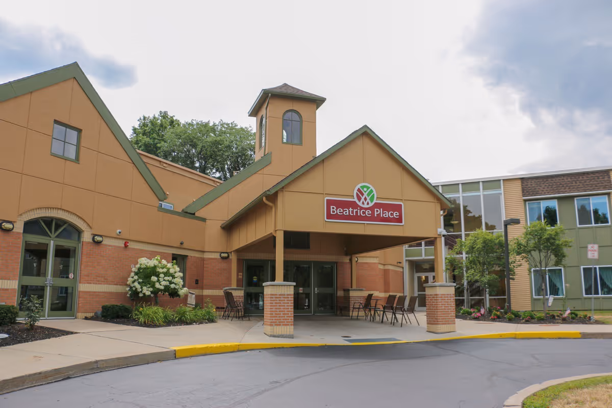 Exterior view of Episcopal SeniorLife - Beatrice Place building entrance with a covered drop-off area, chairs arranged outside, and landscaping including bushes and trees.
