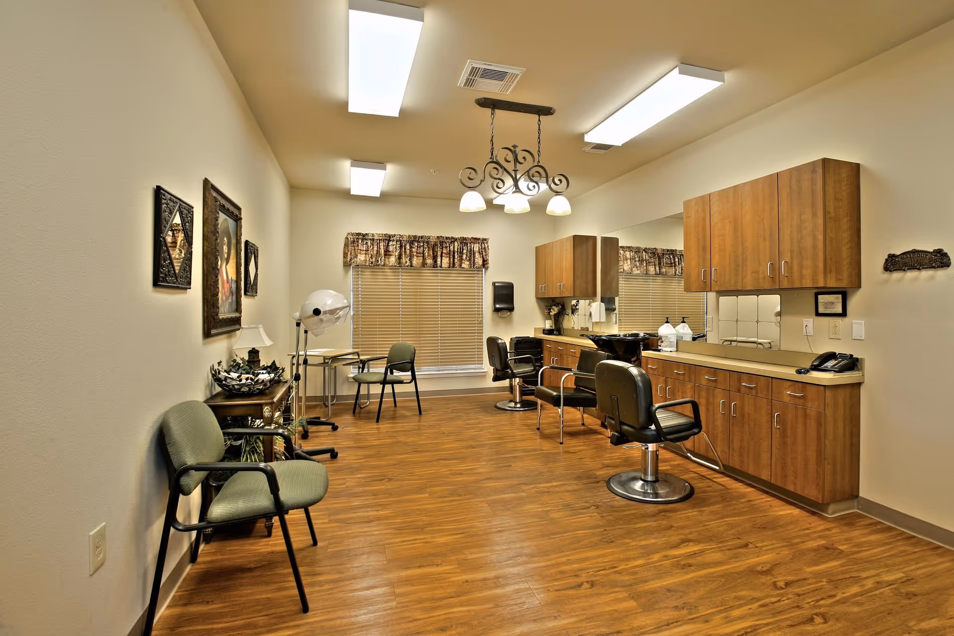Interior of a senior care facility room set up as a hair salon with wooden cabinets, salon chairs, a hair dryer, and framed pictures on the wall. The room has wood flooring, a window with blinds and a valance, and overhead lighting.