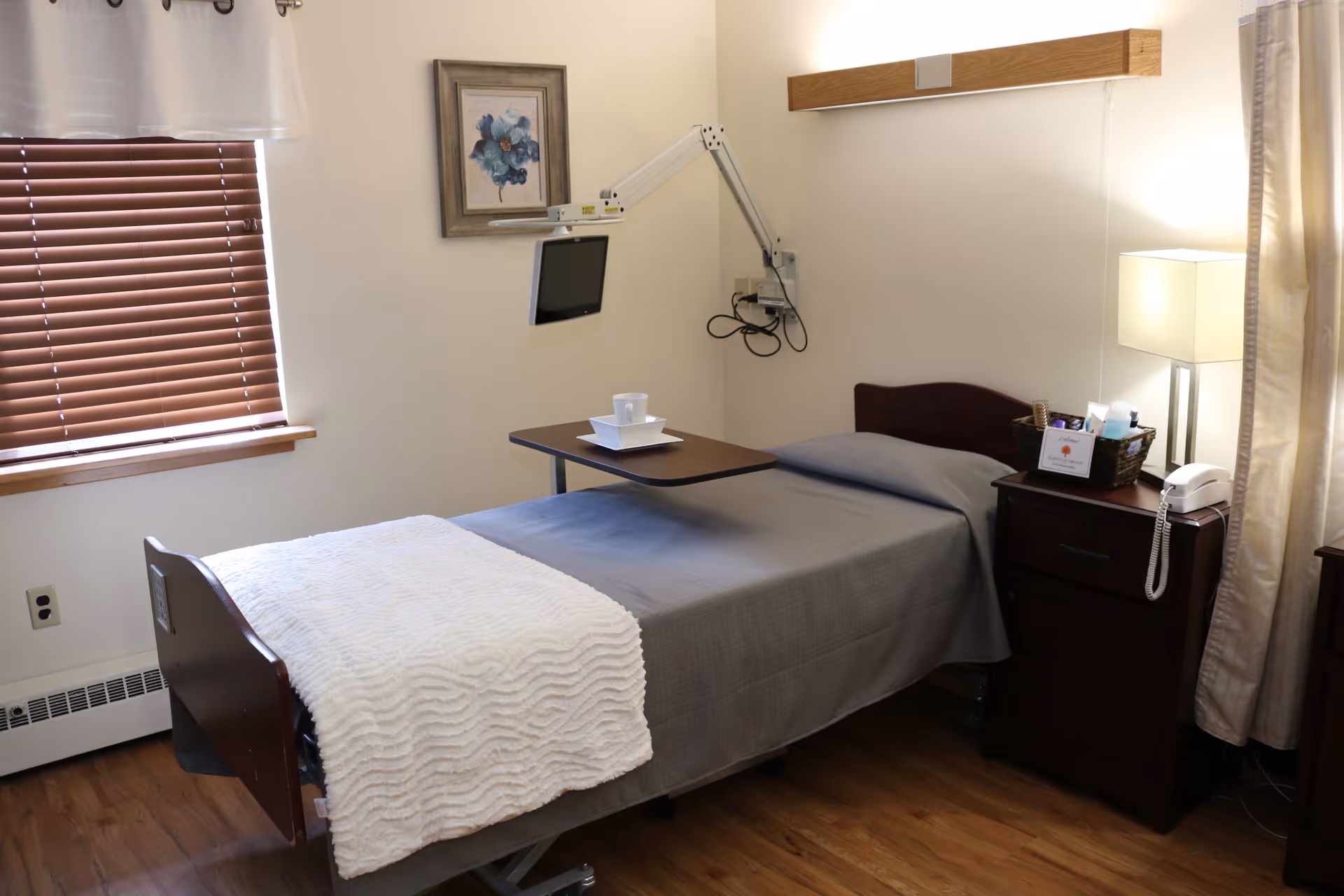 A neatly made single bed in a furnished senior living bedroom with an overbed table, bedside cabinet, lamp, and wall-mounted TV arm.