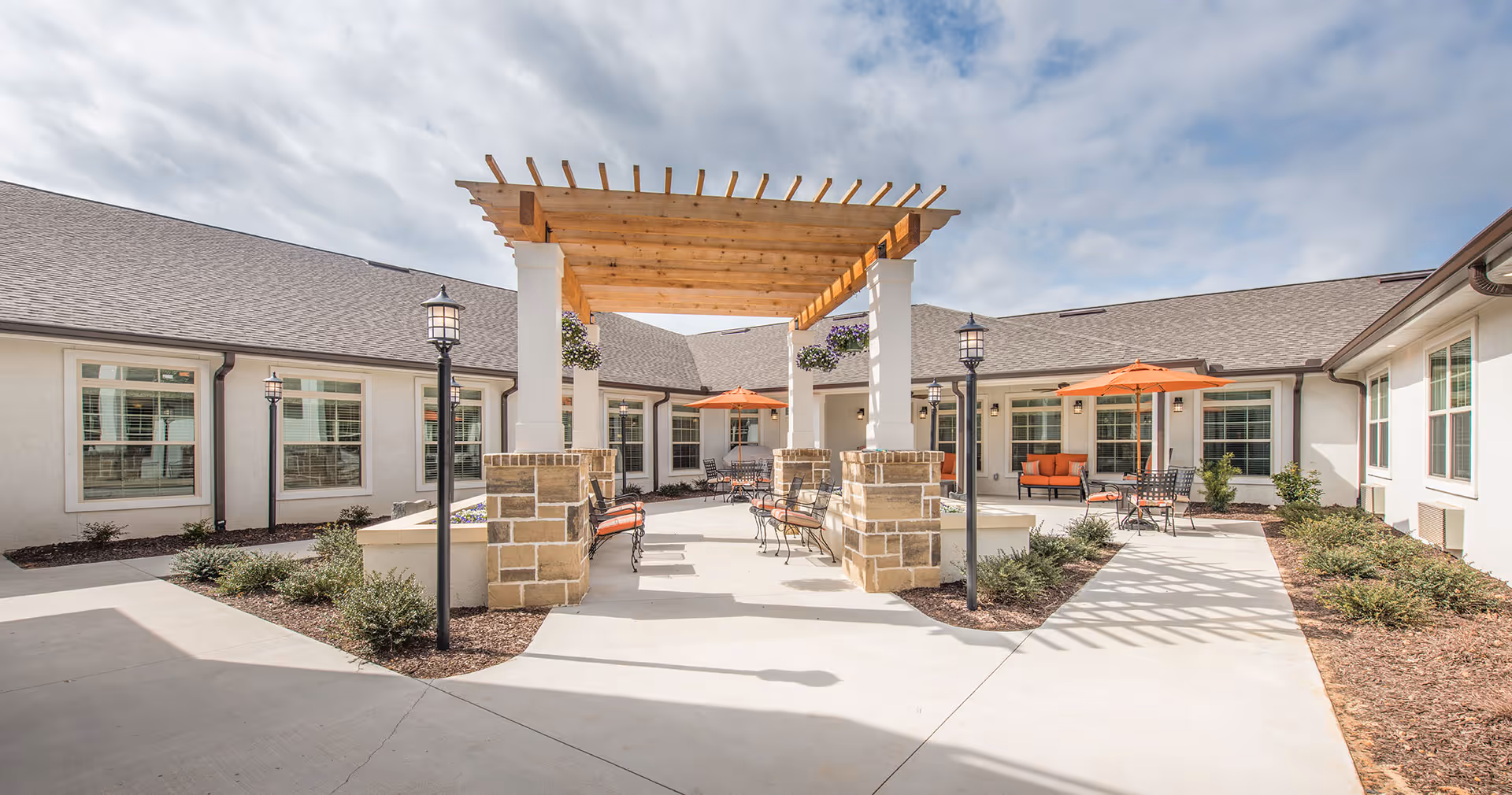 Outdoor courtyard area of Barclay House of Aiken featuring a wooden pergola with hanging flower baskets, several black metal benches with orange cushions, black metal tables with orange umbrellas, surrounded by a white building with multiple windows and landscaped shrubbery under a partly cloudy sky.
