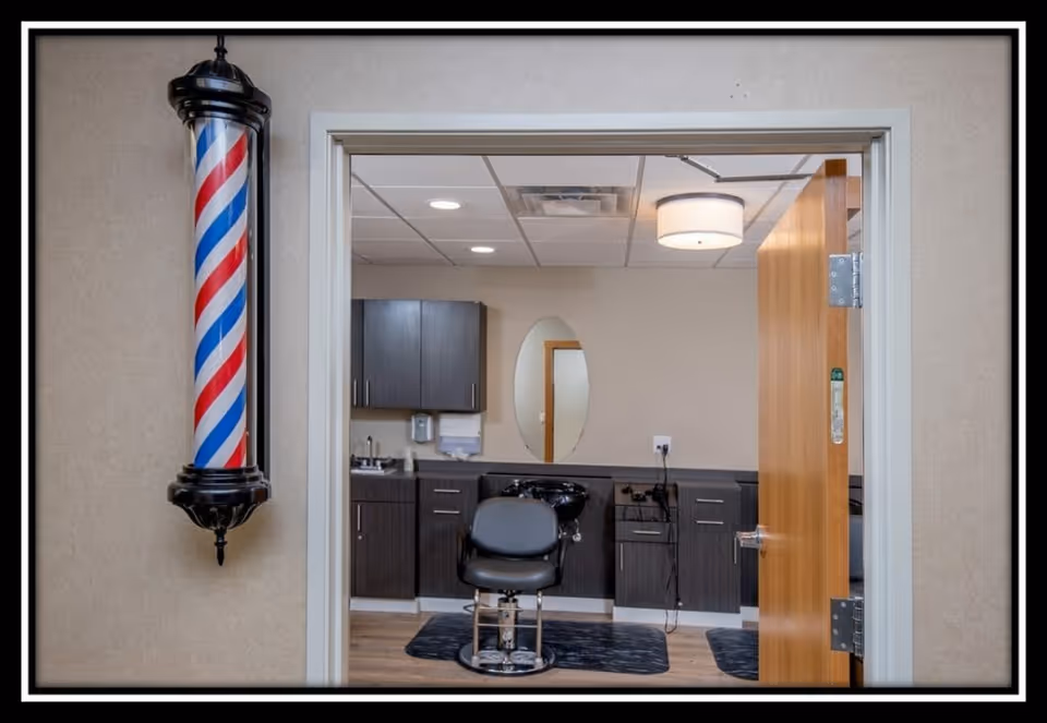 Interior view of a senior care facility's barber or hair salon room. The image shows a black barber chair in front of a counter with dark cabinets, a sink, and an oval mirror on the wall. A wooden door is partially open, and a traditional red, white, and blue barber pole is mounted on the wall outside the room.