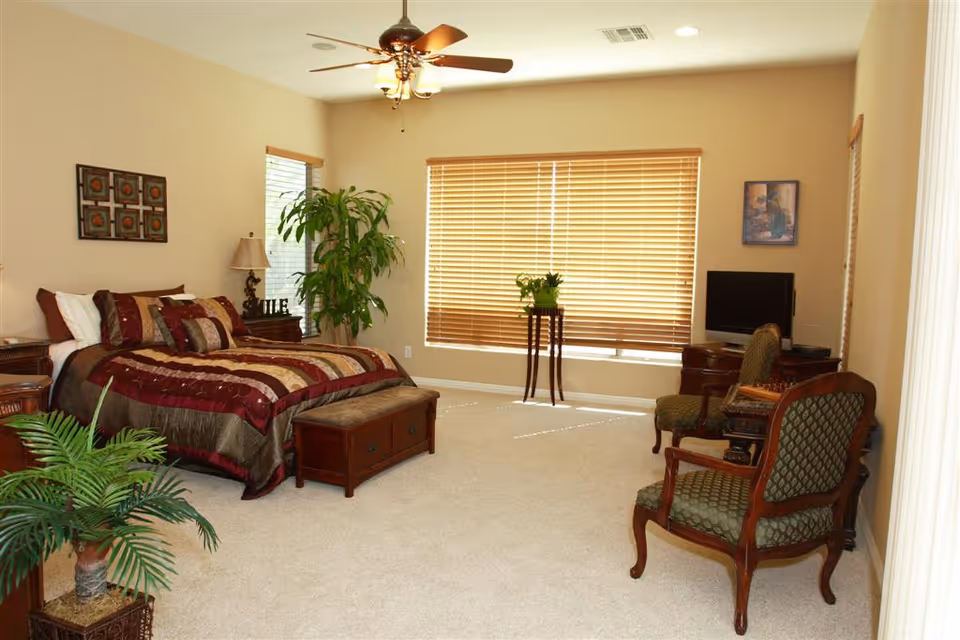 A spacious bedroom with a large bed covered in a red and brown patterned comforter and multiple pillows. There are two wooden nightstands with lamps on either side of the bed. A ceiling fan with lights hangs above. The room has beige walls and carpeted floors. There are two green upholstered chairs with wooden frames, a small wooden table with a plant, a TV on a wooden stand, and large windows with wooden blinds letting in natural light. Several green plants and framed artwork decorate the room.