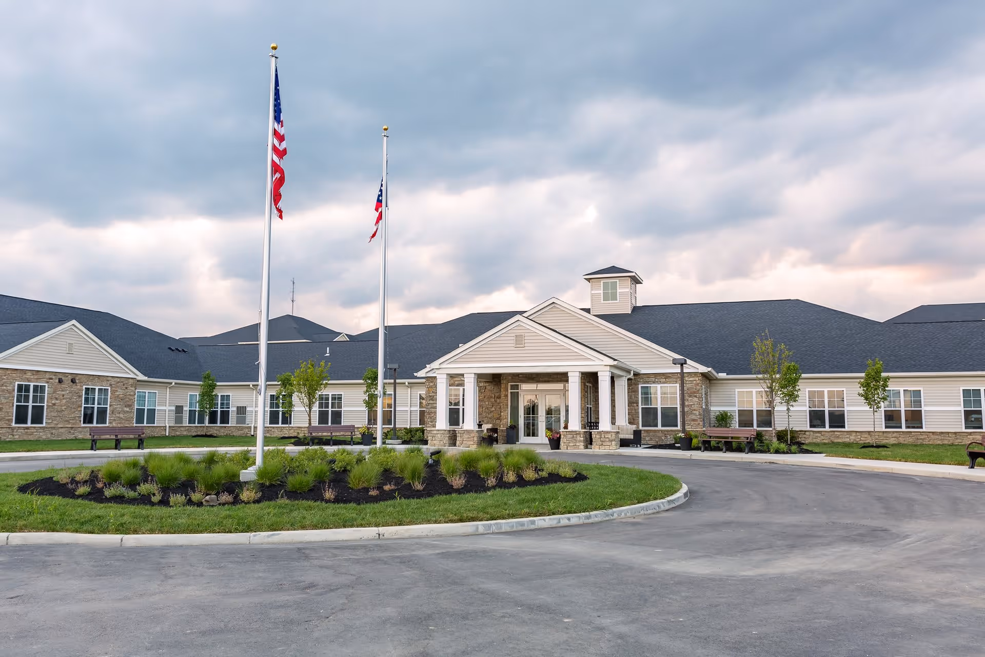 Front exterior view of a single-story senior living facility building with a covered entrance supported by white columns, two flagpoles with American flags, landscaped greenery, and a circular driveway under a cloudy sky.