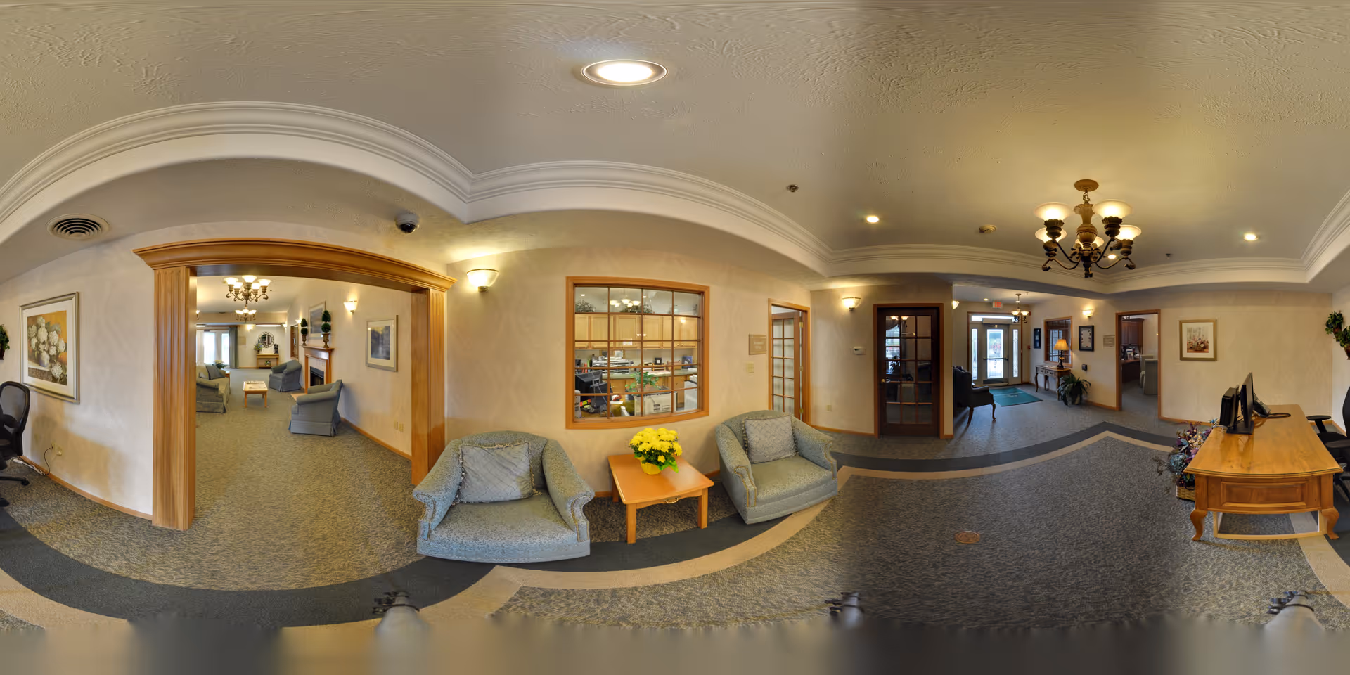 Panoramic interior of a senior living residence lobby showing seating areas, a reception desk, chandeliers, and hallways.