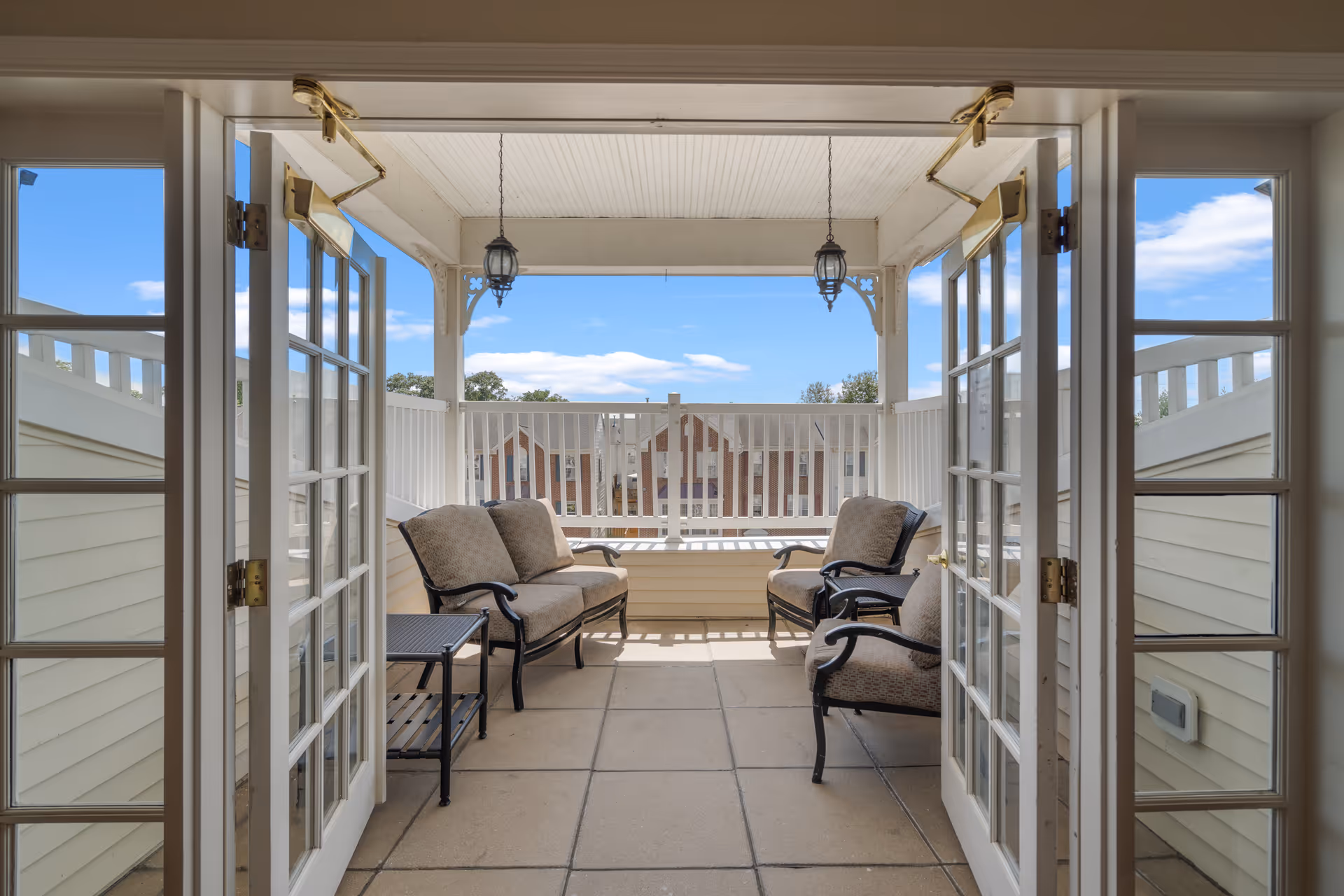 View through open double glass doors onto a covered outdoor balcony with cushioned seating including a loveseat and two armchairs, a small side table, hanging lantern-style lights, and a white railing overlooking neighboring rooftops under a blue sky with some clouds.