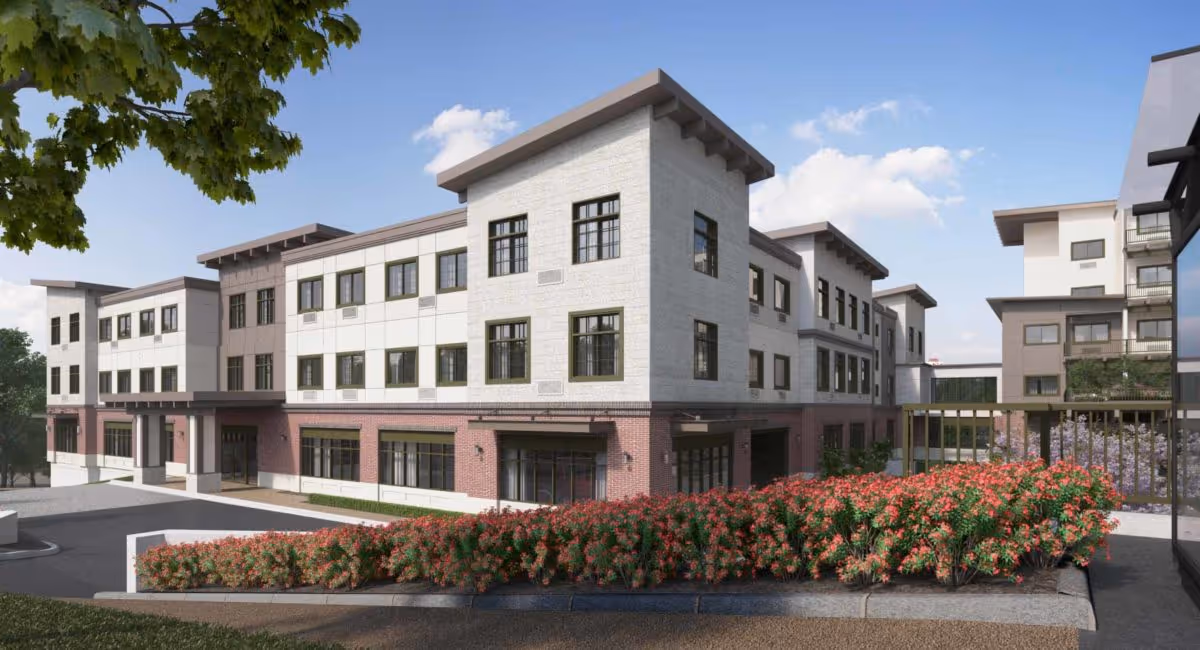 Exterior view of a modern three-story assisted living community building with a combination of white and brick facade, large windows, and landscaped bushes with red flowers in the foreground under a partly cloudy sky.