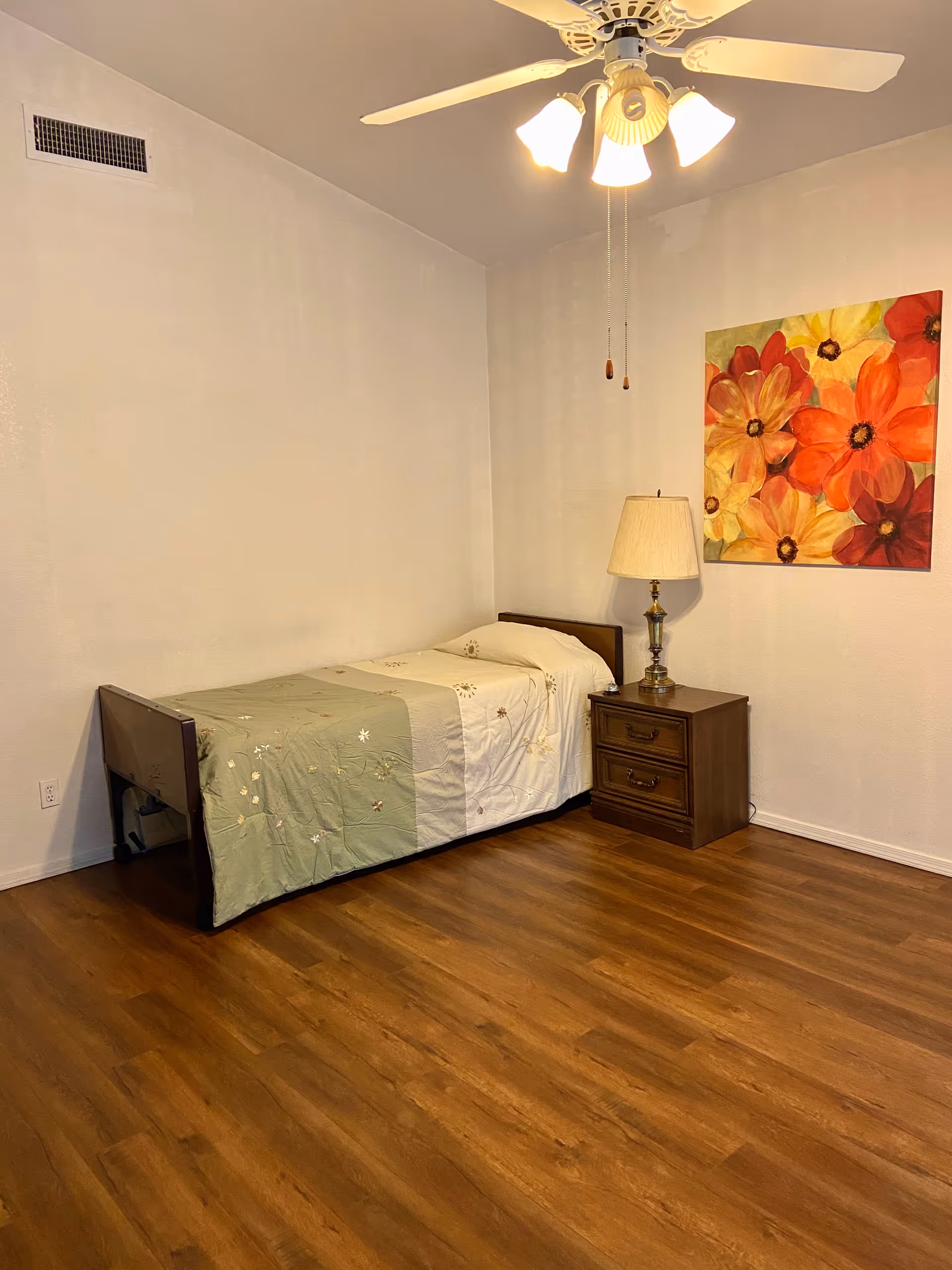 A simple bedroom with a single bed covered with a green and beige floral bedspread, a wooden nightstand with a lamp, a colorful floral painting on the wall, and a ceiling fan with lights above. The room has wooden flooring and plain light-colored walls.