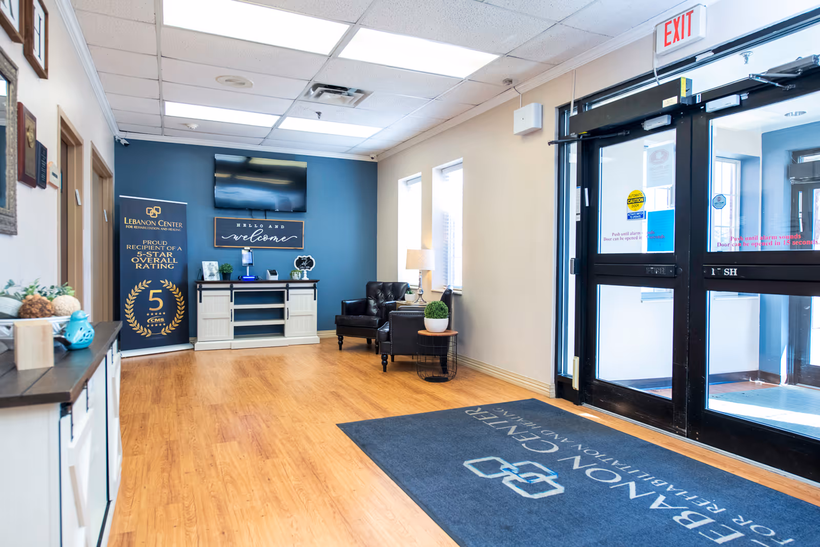 Bright and welcoming entrance area of Lebanon Center for Rehabilitation and Healing featuring wooden flooring, a dark blue accent wall with a mounted TV and a welcome sign, a banner highlighting a 5-star overall rating, a black leather chair with a side table and lamp, and glass double doors with exit signs and caution stickers. A branded floor mat is placed near the entrance.