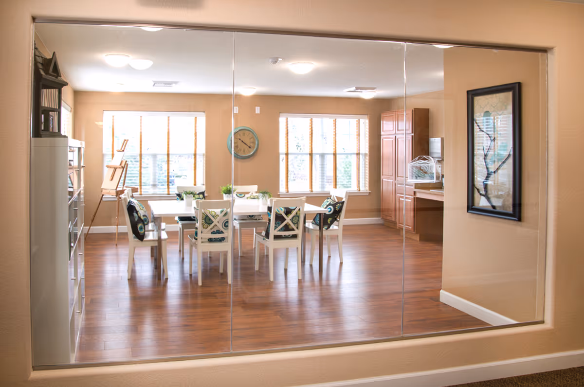 View through a large interior window into a bright dining room with a white table and six chairs, some with patterned cushions. The room has wooden floors, beige walls, two large windows with blinds, a wall clock, a framed picture, and wooden cabinets in the corner.