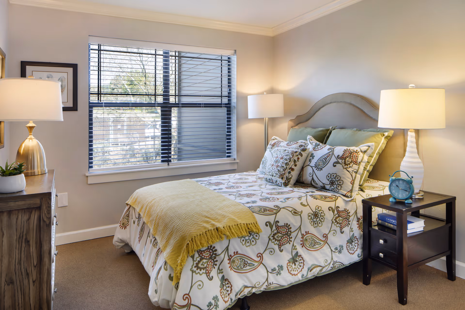 A cozy bedroom with a bed featuring floral patterned bedding and multiple pillows. There is a yellow throw blanket at the foot of the bed. On the right side of the bed is a dark wooden nightstand with a white lamp, a blue alarm clock, and some books. On the left side, there is a wooden dresser with a gold lamp and a small potted plant. A large window with blinds lets in natural light, and the walls are painted a soft beige color.