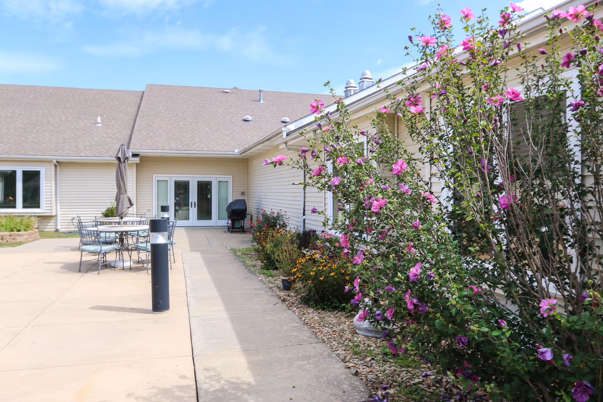 Outdoor patio area at Vintage Park at Baldwin City featuring a concrete walkway, metal tables and chairs with an umbrella, a barbecue grill, and a garden bed with blooming pink flowers along the building's exterior wall under a clear blue sky.