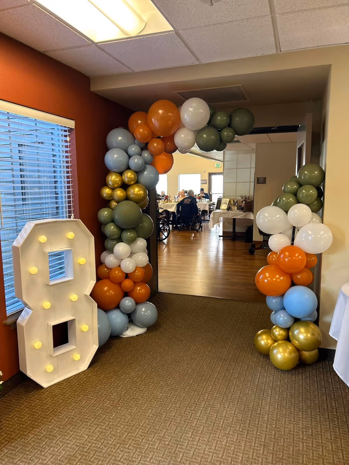 An indoor hallway decorated with a colorful balloon arch in shades of orange, green, white, blue, and gold. To the left of the arch is a large illuminated number 8. Beyond the arch, a dining area is visible with elderly people seated at tables.