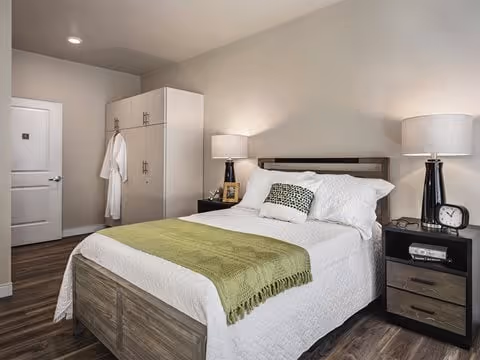 Bedroom with a neatly made bed, two bedside tables with lamps, a wardrobe, and wood flooring.