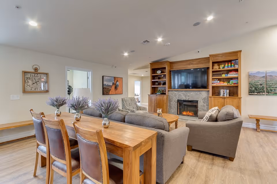 A cozy living room area with a wooden table and four leather chairs in the foreground, decorated with three vases of purple flowers. Behind the table are two gray sofas and an armchair arranged around a wooden coffee table. A stone fireplace with a mounted flat-screen TV above it is flanked by wooden built-in shelves filled with books and games. The room has light-colored walls, wood flooring, and recessed ceiling lights. A large clock and framed pictures hang on the walls.