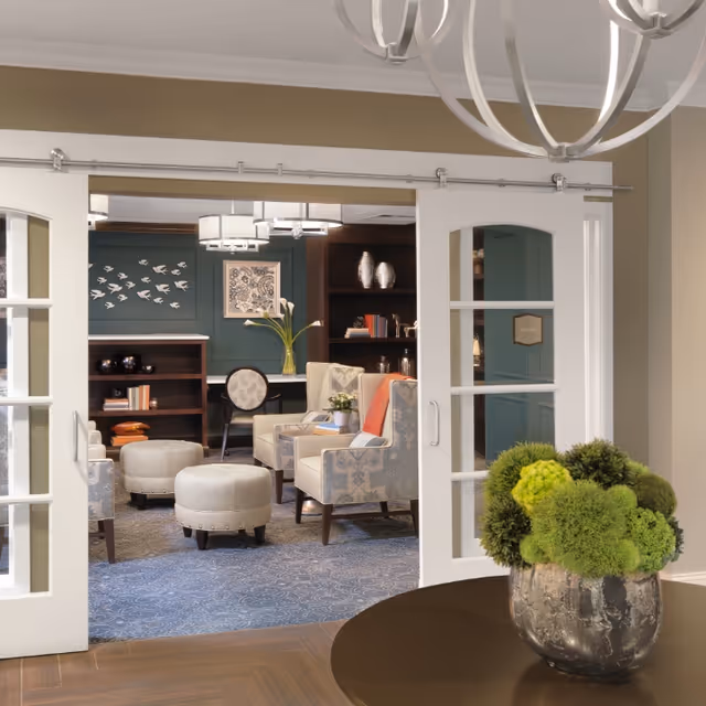 A cozy living room area viewed through white sliding glass doors. The room features patterned armchairs, round ottomans, a bookshelf with decorative items, and a wall adorned with bird decorations and framed artwork. A round table with a green floral arrangement is in the foreground, and a modern chandelier hangs from the ceiling.