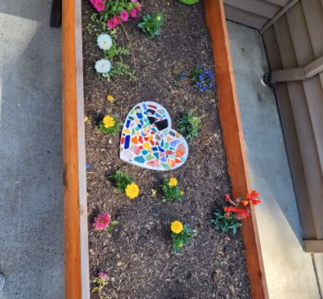 A raised garden bed with various colorful flowers planted in soil. In the center of the garden bed is a heart-shaped mosaic decoration made of multicolored pieces. The garden bed is bordered by wooden planks and is situated next to a concrete walkway and a building wall.