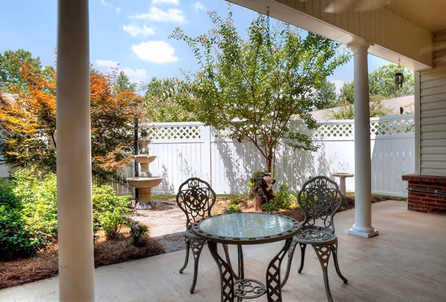 Covered patio area with a round glass-top metal table and three matching chairs. The patio overlooks a garden with a white lattice fence, a small tree, bushes, and a tiered water fountain. The sky is blue with some clouds.