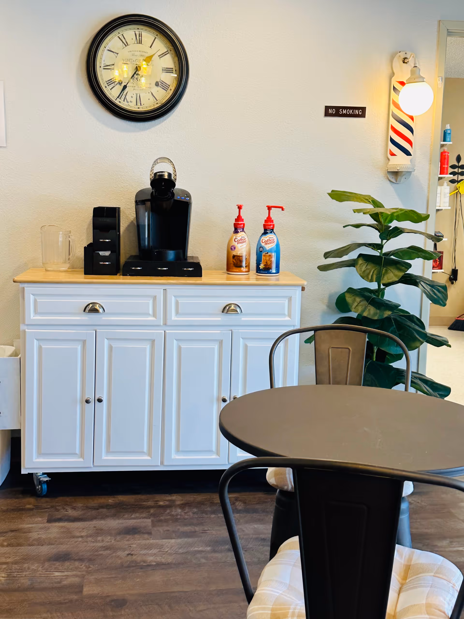 A coffee station with a black coffee maker, two bottles of Coffee-mate creamer, a clear pitcher, and a black organizer on a white cabinet with a wooden countertop. Above the cabinet is a round wall clock with Roman numerals and a small 'No Smoking' sign on the wall. To the right, there is a green potted plant and a wall-mounted light fixture with a barber pole design. In the foreground, there is a round black table with black metal chairs that have beige cushioned seats.