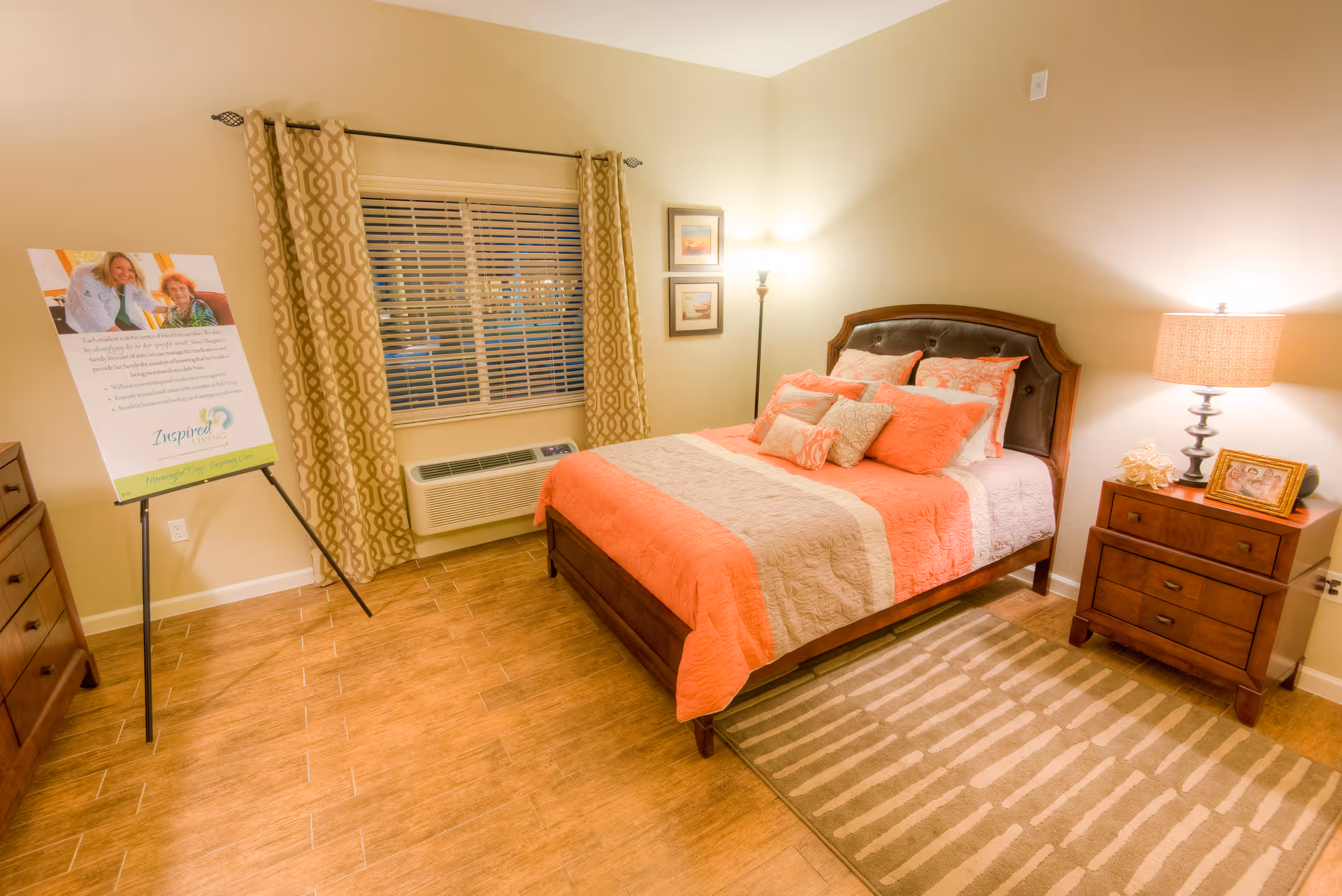 A cozy bedroom with a wooden bed featuring coral and beige bedding and multiple pillows. There is a wooden nightstand with a lamp and a framed photo on it. A floor lamp stands in the corner next to two framed pictures on the wall. The window has patterned curtains and a white air conditioning unit below it. A wooden dresser is partially visible on the left side, and a sign on a tripod stand is placed near the window. The floor is wooden with a beige and white striped rug beside the bed.