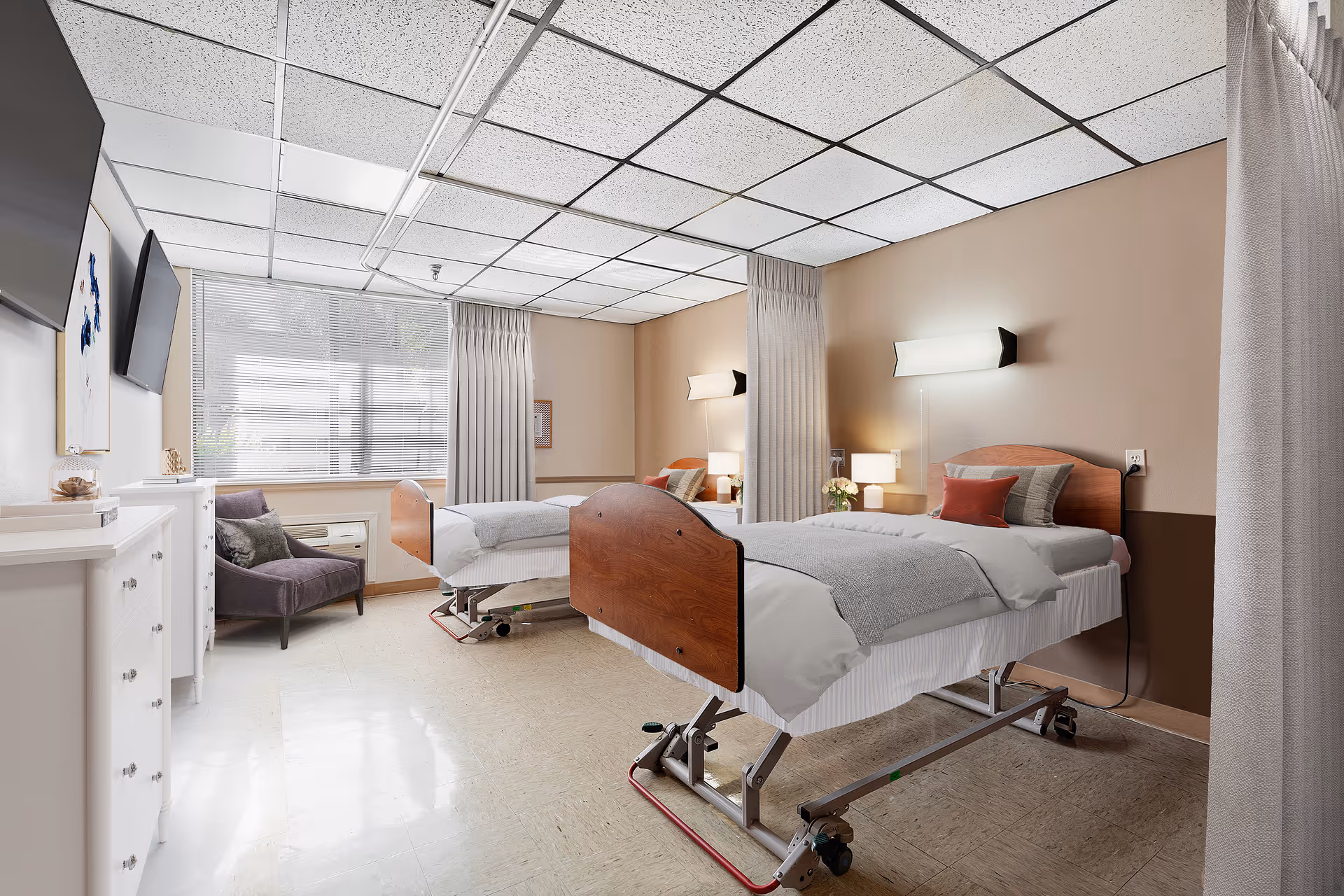 A bright and clean nursing facility room with two hospital beds, each with wooden headboards and footboards, white bedding, and gray blankets. The room has beige walls with a darker brown lower section, a large window with blinds and curtains, a purple armchair, a white dresser, wall-mounted lights above each bed, and a flat-screen TV mounted on the wall.