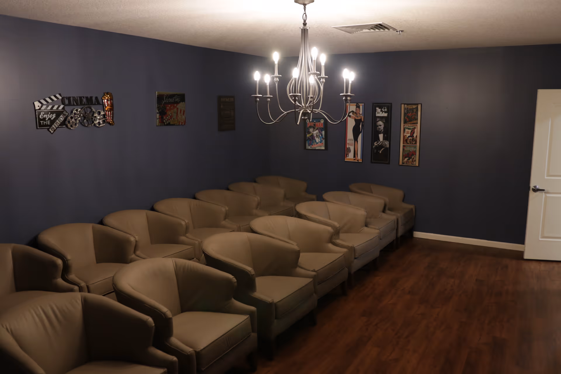 A small screening room with multiple beige armchairs arranged in rows, a chandelier overhead, dark blue walls decorated with movie posters, and a wood floor.