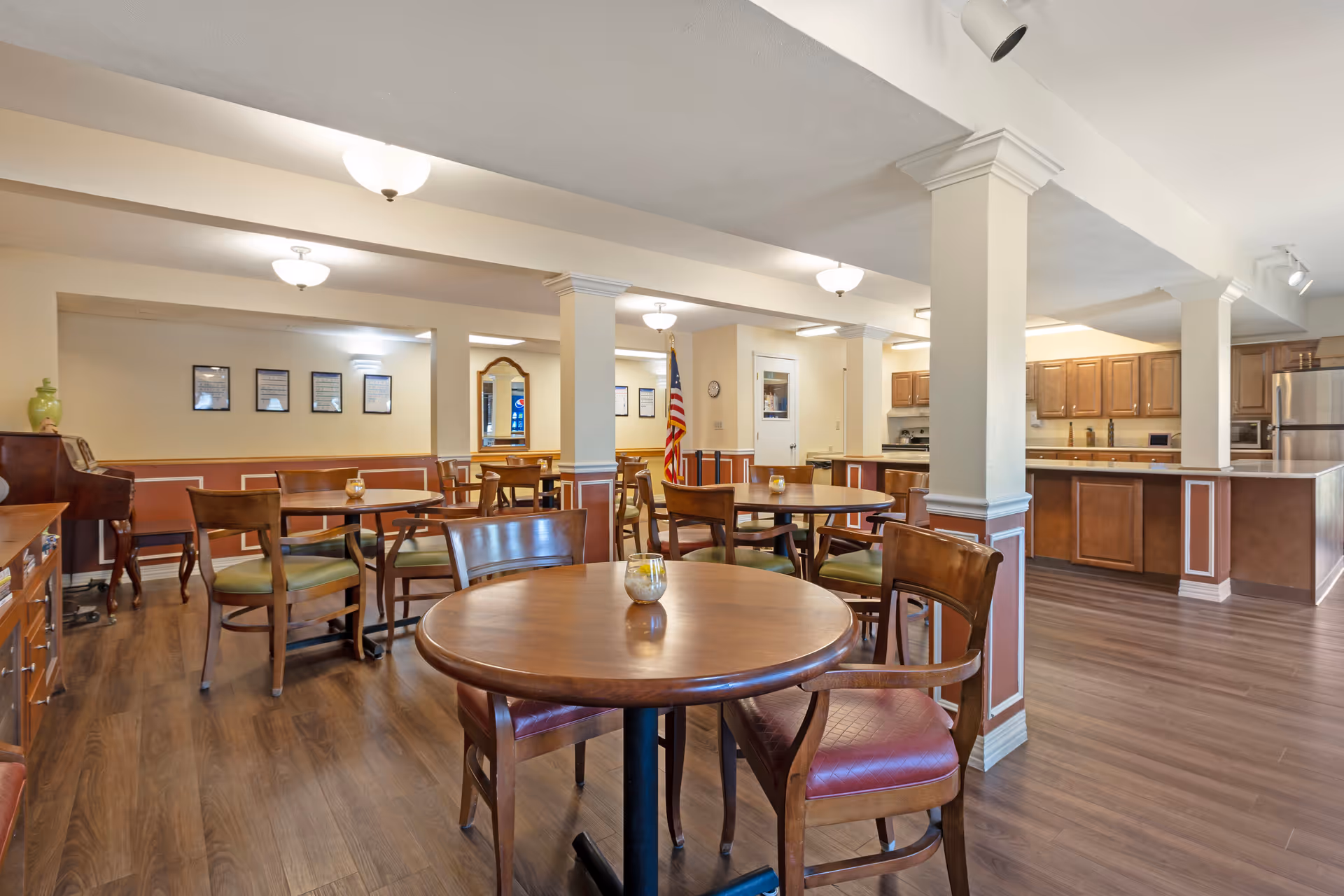 A dining area in a senior living facility featuring round wooden tables with chairs arranged around them. The room has wooden flooring, beige walls with white trim, and several ceiling lights. In the background, there is a kitchen area with wooden cabinets and stainless steel appliances. An American flag is visible near the back wall, along with framed pictures and a mirror.