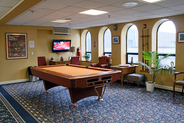 A carpeted common room with a pool table, upholstered chairs, a wall-mounted TV, and arched windows.
