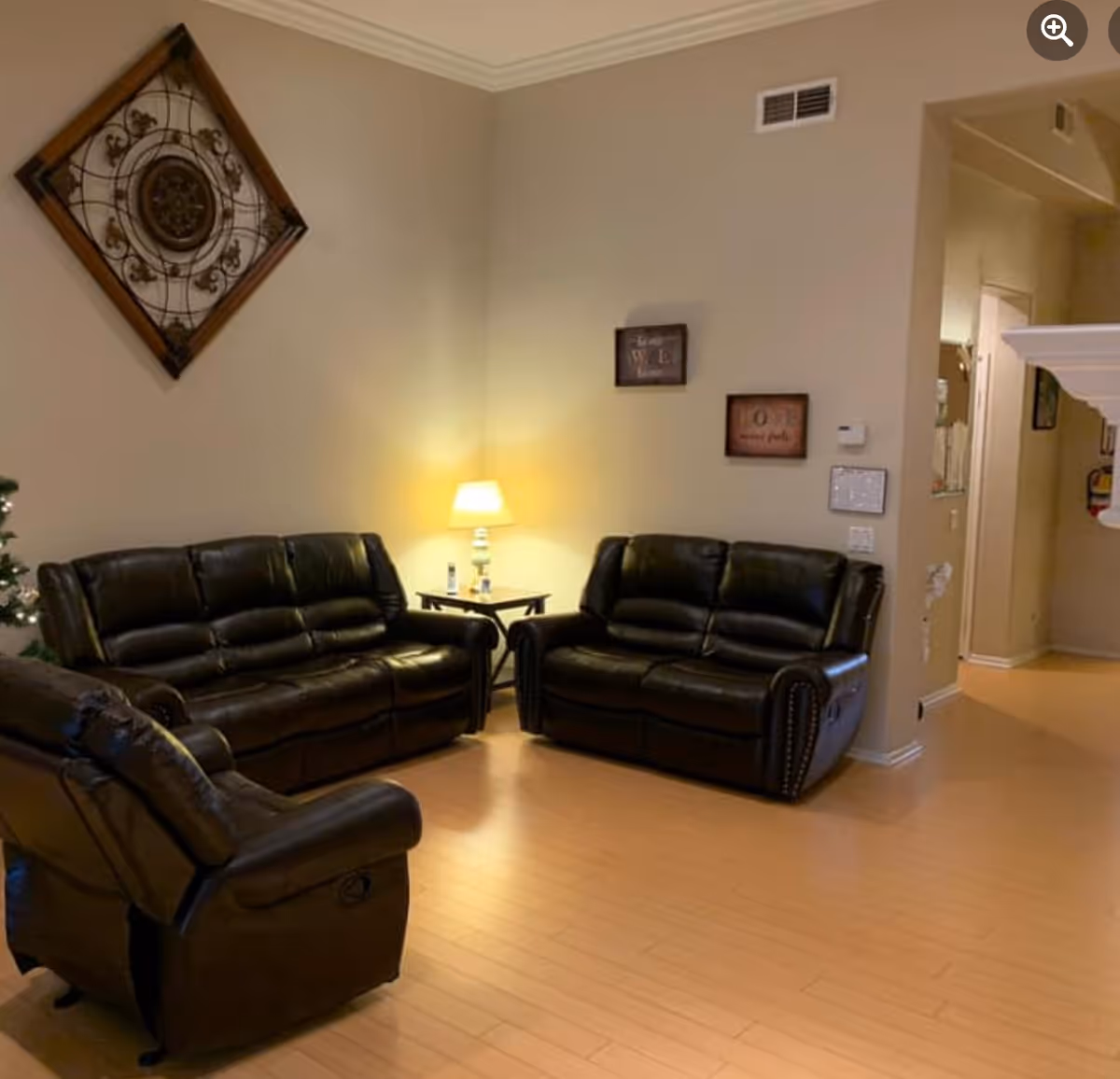 A cozy living room with three dark brown leather recliner sofas arranged around a corner. A small table with a lit lamp is placed between two of the sofas. The walls are beige with decorative wall art, and the floor is light wood. There is an open doorway leading to another room.