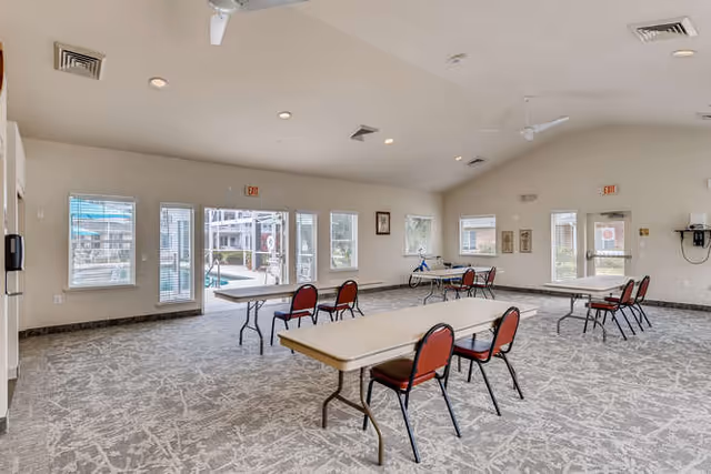 Large bright community room with folding tables, red chairs, and windows/doors overlooking an outdoor pool area.