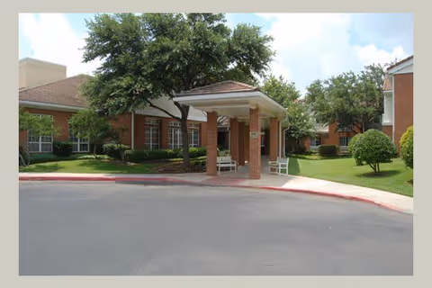 Entrance area of a senior living facility with a covered drop-off zone supported by brick columns, surrounded by well-maintained green lawns, trees, and shrubs. The building has a brick exterior and multiple windows, with a clear blue sky in the background.