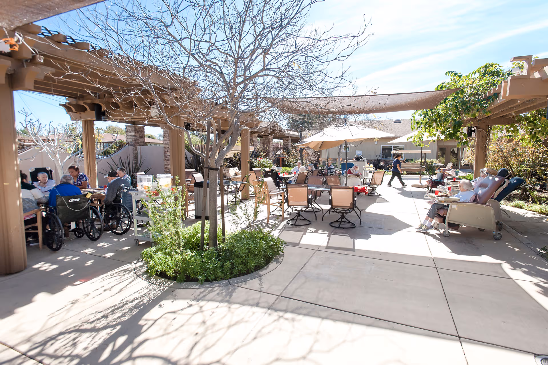 Sunny outdoor courtyard with pergolas, tables, umbrellas and residents seated in chairs and wheelchairs.