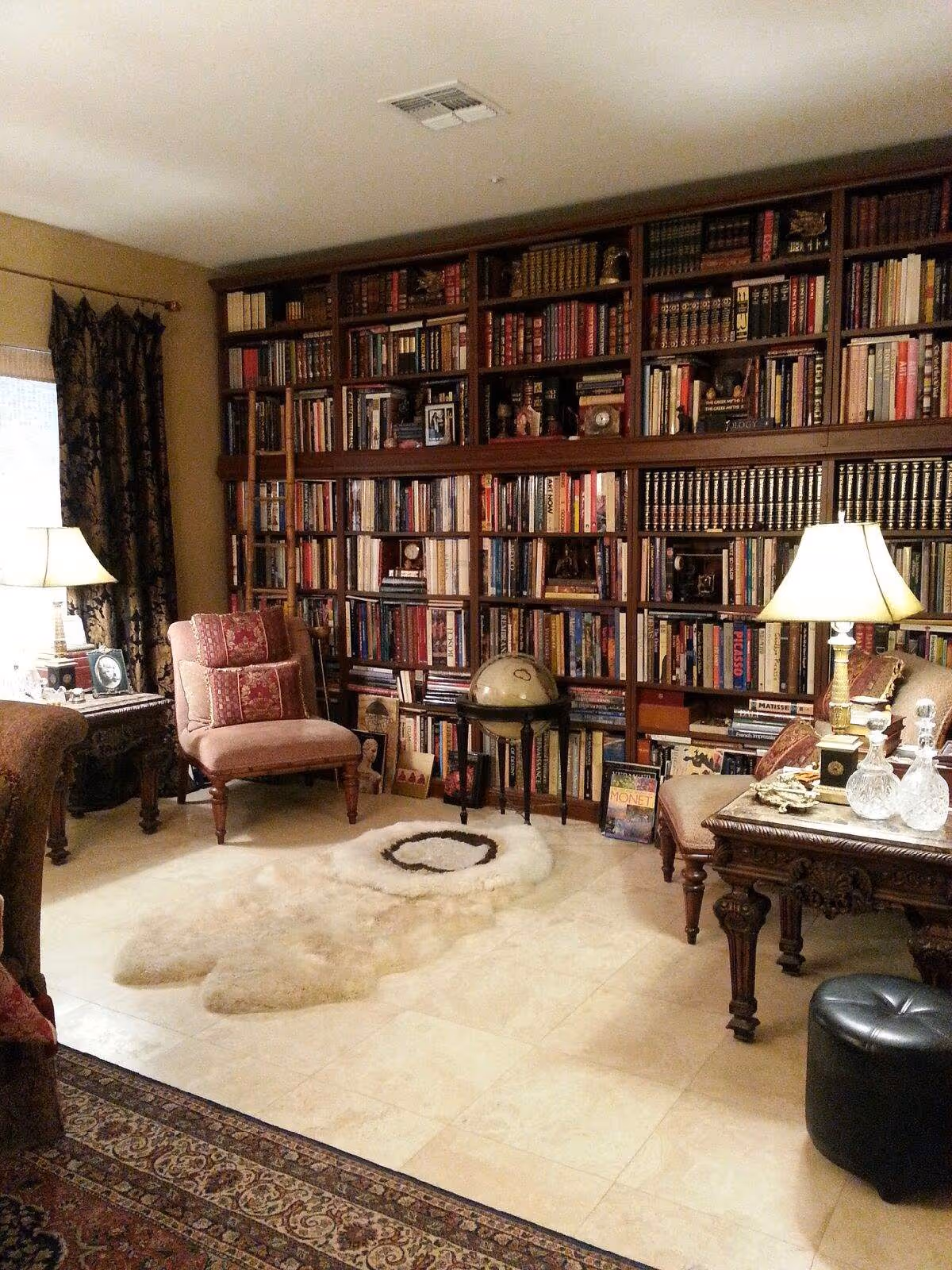 Cozy living room library with a wall of bookshelves, armchairs, lamps, a globe, and rugs on a tiled floor.