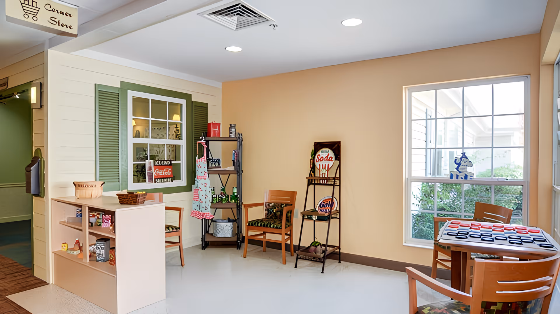 Interior corner store area in a senior living facility with a small counter displaying snacks, a shelving unit with aprons and decorative items, two wooden chairs, and a table with a checkers game near a large window showing outdoor greenery.
