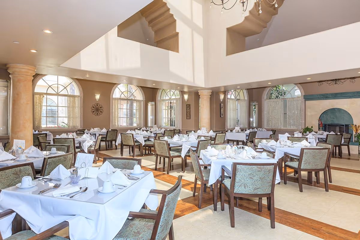 A bright and spacious dining room with multiple tables covered in white tablecloths, each set with white napkins, cups, and silverware. The room features large arched windows allowing natural light to fill the space, beige walls, decorative columns, and a high ceiling with a chandelier.