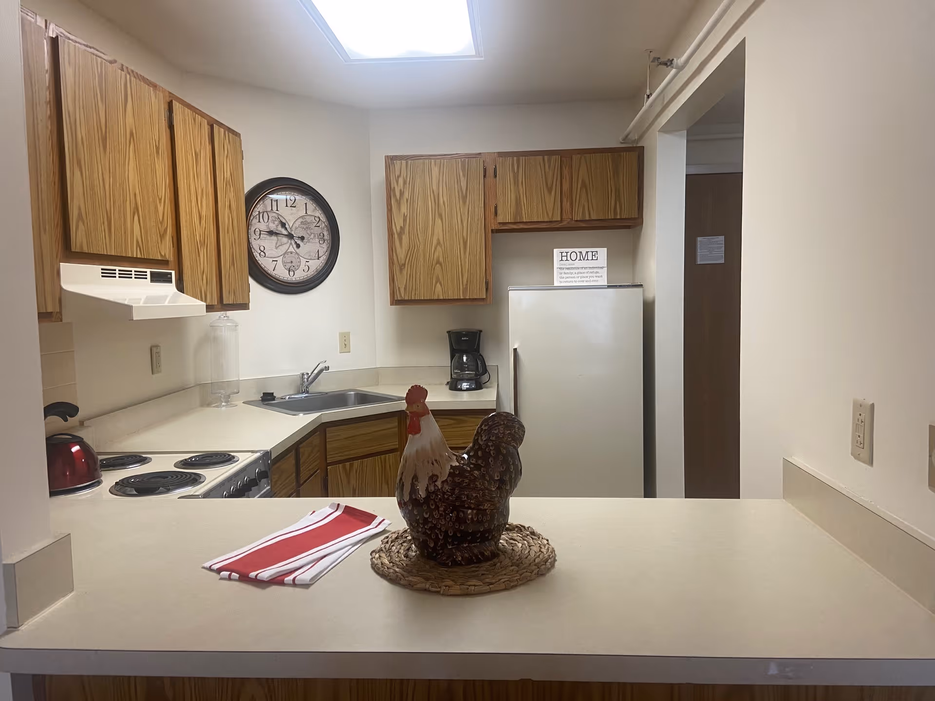 A small kitchen with wooden cabinets, a white countertop, a stove with a red kettle, a sink, a coffee maker, and a white refrigerator. A decorative ceramic rooster sits on a woven mat on the countertop along with a red and white striped cloth. A large round wall clock is mounted on the wall above the sink. The kitchen is lit by a skylight.