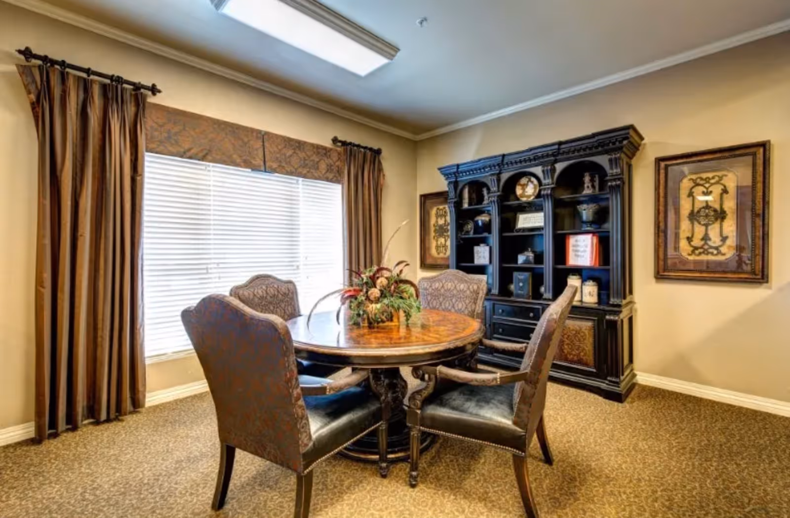 A cozy interior room with a round wooden table surrounded by four upholstered chairs. The table has a floral centerpiece. Behind the table is a large dark wooden cabinet with shelves displaying decorative items. The room has beige walls, carpeted floor, a large window with closed blinds and brown curtains, and framed artwork on the walls.