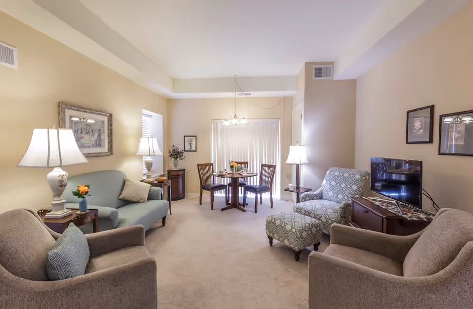 Bright furnished living room with sofas, armchairs, lamps, a TV and a small dining table near sliding blinds.