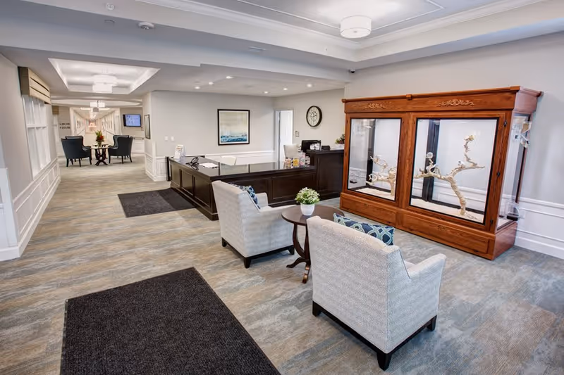 A bright and spacious reception area in a senior living facility with a dark wood reception desk, two light-colored armchairs with patterned cushions around a small round table with a plant, and a large wooden display case containing decorative branches. The hallway extends into the background with additional seating and wall art.