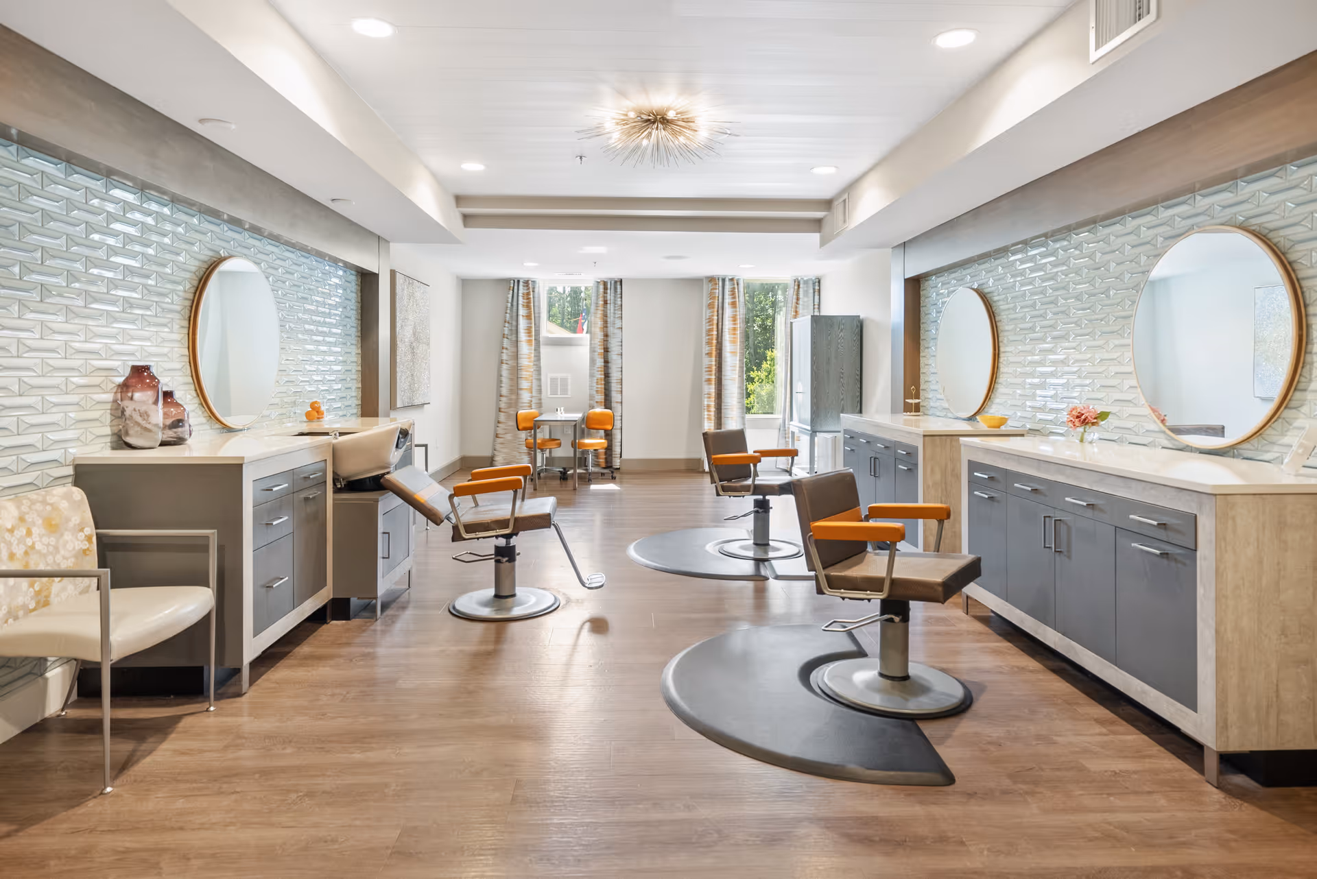 A bright and modern salon area with three styling chairs positioned in front of large round mirrors mounted on a tiled wall. The room features wood flooring, gray cabinetry with white countertops, and decorative vases and flowers. In the background, there is a small table with two orange chairs near windows with patterned curtains.