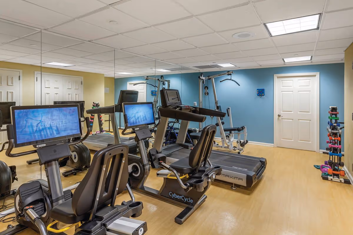 A fitness room with exercise equipment including two recumbent stationary bikes with screens, a treadmill, and a multi-function weight machine. The room has light wood flooring, a blue accent wall, a mirrored wall, and a rack of colorful dumbbells in the corner.