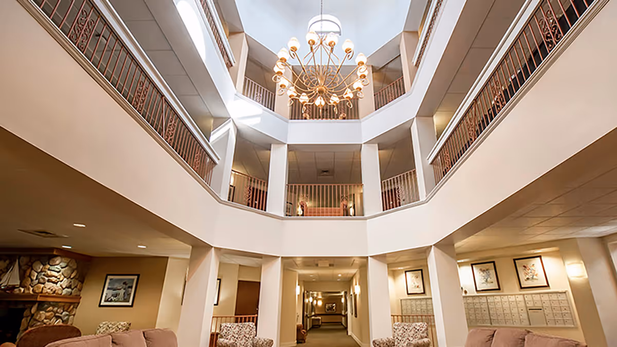 Interior view of a senior living facility lobby with a high ceiling, a large chandelier, multiple seating areas with armchairs and sofas, and a stone fireplace on the left side. The lobby has two upper floors with railings overlooking the central area and mailboxes on the right wall.