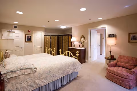 A cozy bedroom in a nursing center featuring a large bed with a floral bedspread, a wooden dresser with a lamp and decorative items, a comfortable upholstered armchair, and soft lighting. The room has beige walls and carpet, with multiple doors leading to other areas.