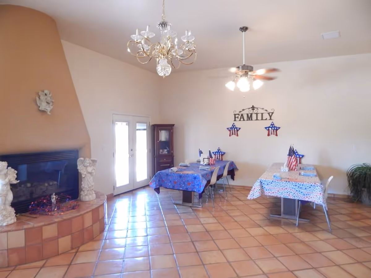 A spacious dining area with two tables covered in patriotic-themed tablecloths and small American flags as centerpieces. The room features a tiled floor, a large fireplace with decorative sculptures, a chandelier, ceiling fan with lights, and a wall decoration that reads 'FAMILY' with star-shaped patriotic ornaments. French doors allow natural light into the room.
